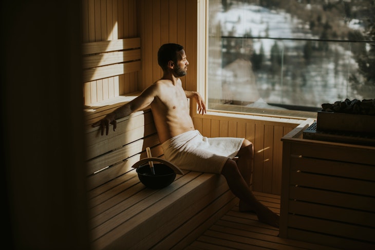 Handsome young man relaxing in the sauna and watching winter forest through the window