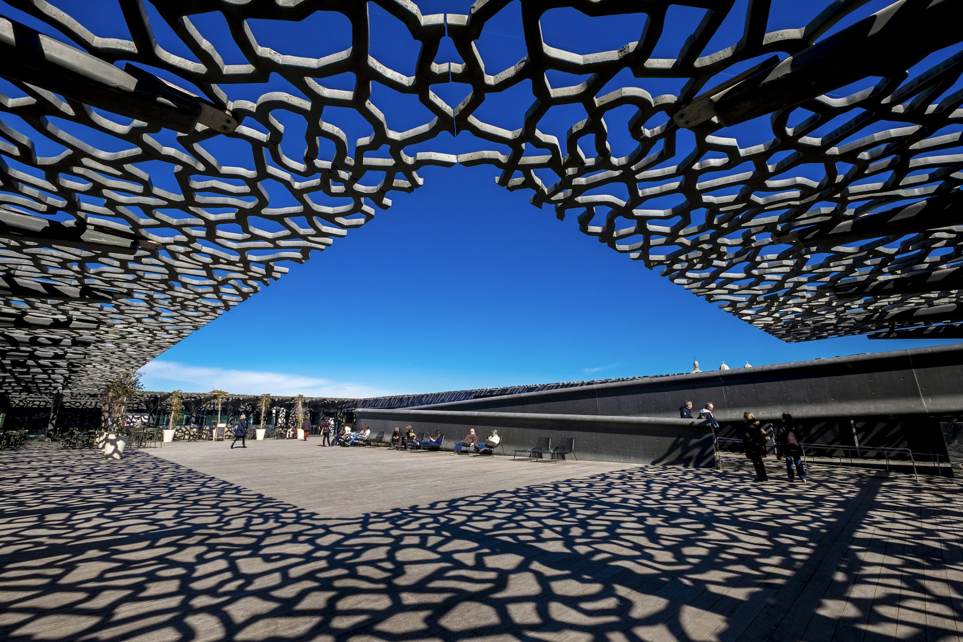 A lattice of metal forms part of the roof of a museum building, creating a network of shadows on the ground in an open square full of visitors