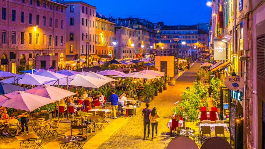 Night view of a square full of restaurants at Port Vieux in Marseille. People are sat at tables under large white umbrellas