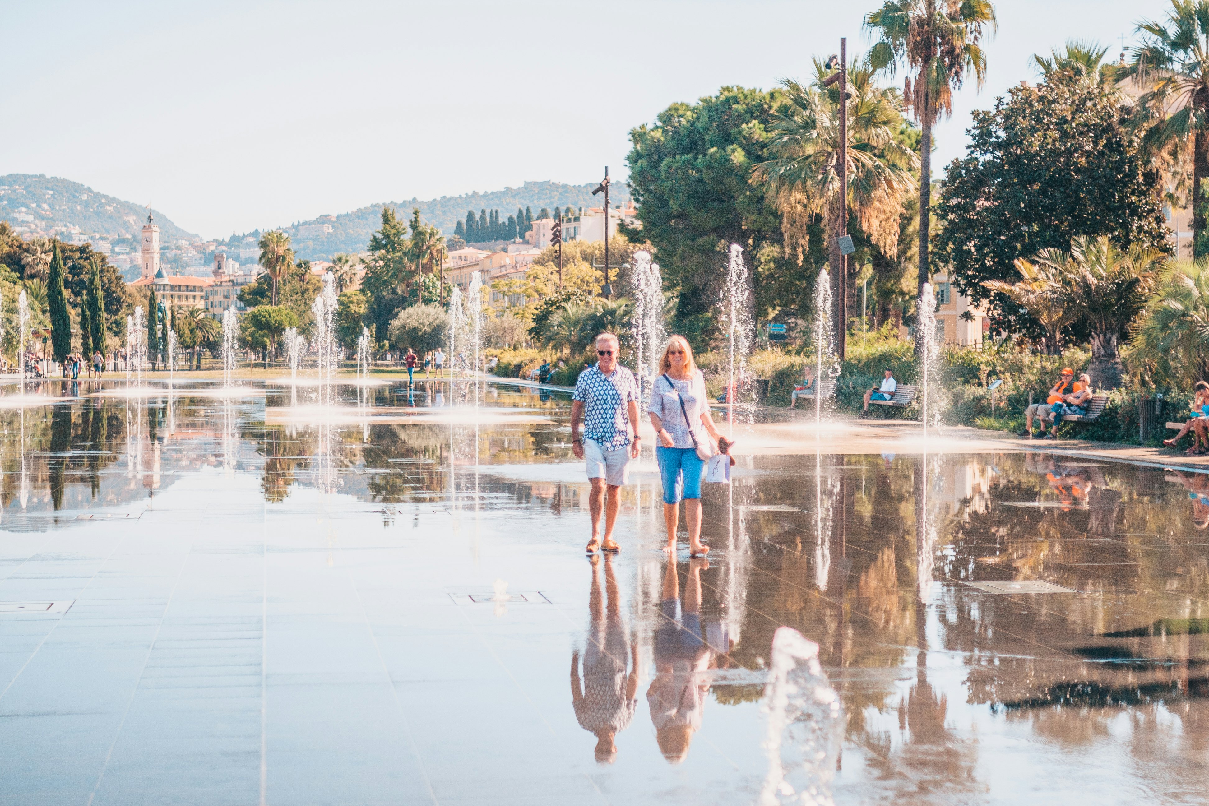 A middle-aged holidaying couple stroll through a square with water jets and fountains shooting out of the sidewalk in a palm-fringed square