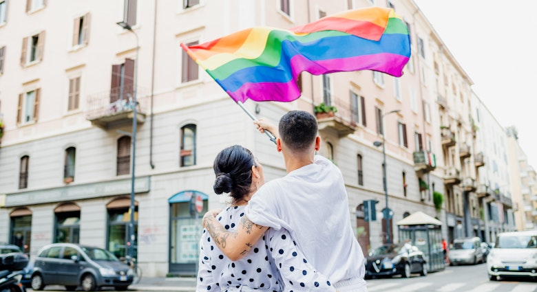Rearview of young lesbian couple standing on a street, waving a rainbow flag