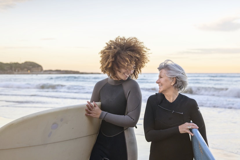 Vibrant senior woman and young mixed race woman surfing together at the beach. The multi-generational female friends are walking towards the camera out of the ocean. They are carrying their surfboards and are talking to each other happily. Both individuals are wearing wetsuits. Healthy lifestyle, hobbies, travel, and surfing concepts.