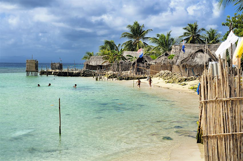 Las aguas azules lamen una playa con edificios con techo de paja en Nalunega, Islas San Blas, Panamá 