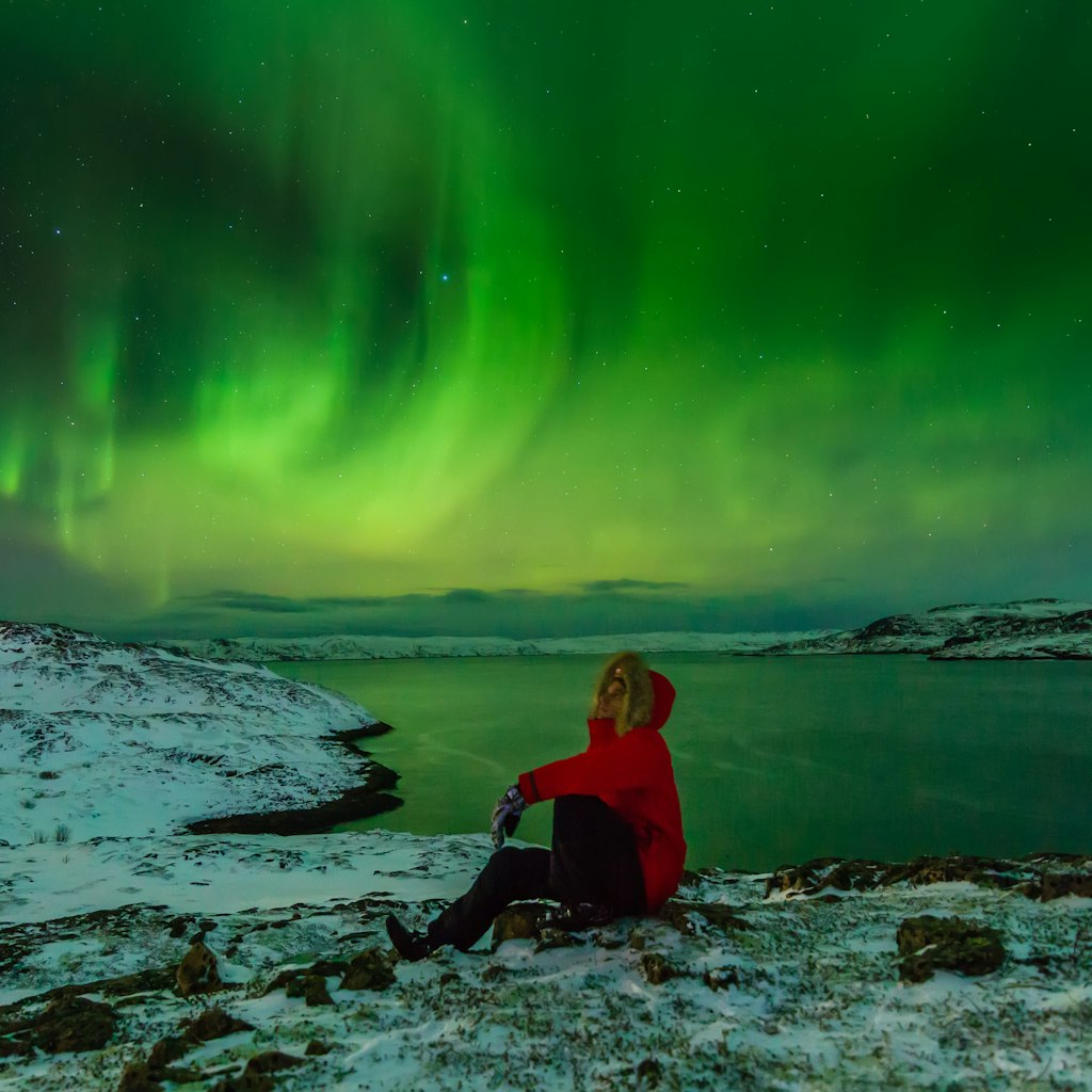 A man sits on the snow with the Northern Lights in the sky on the North Sea shore.