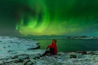 A man sits on the snow with the Northern Lights in the sky on the North Sea shore.