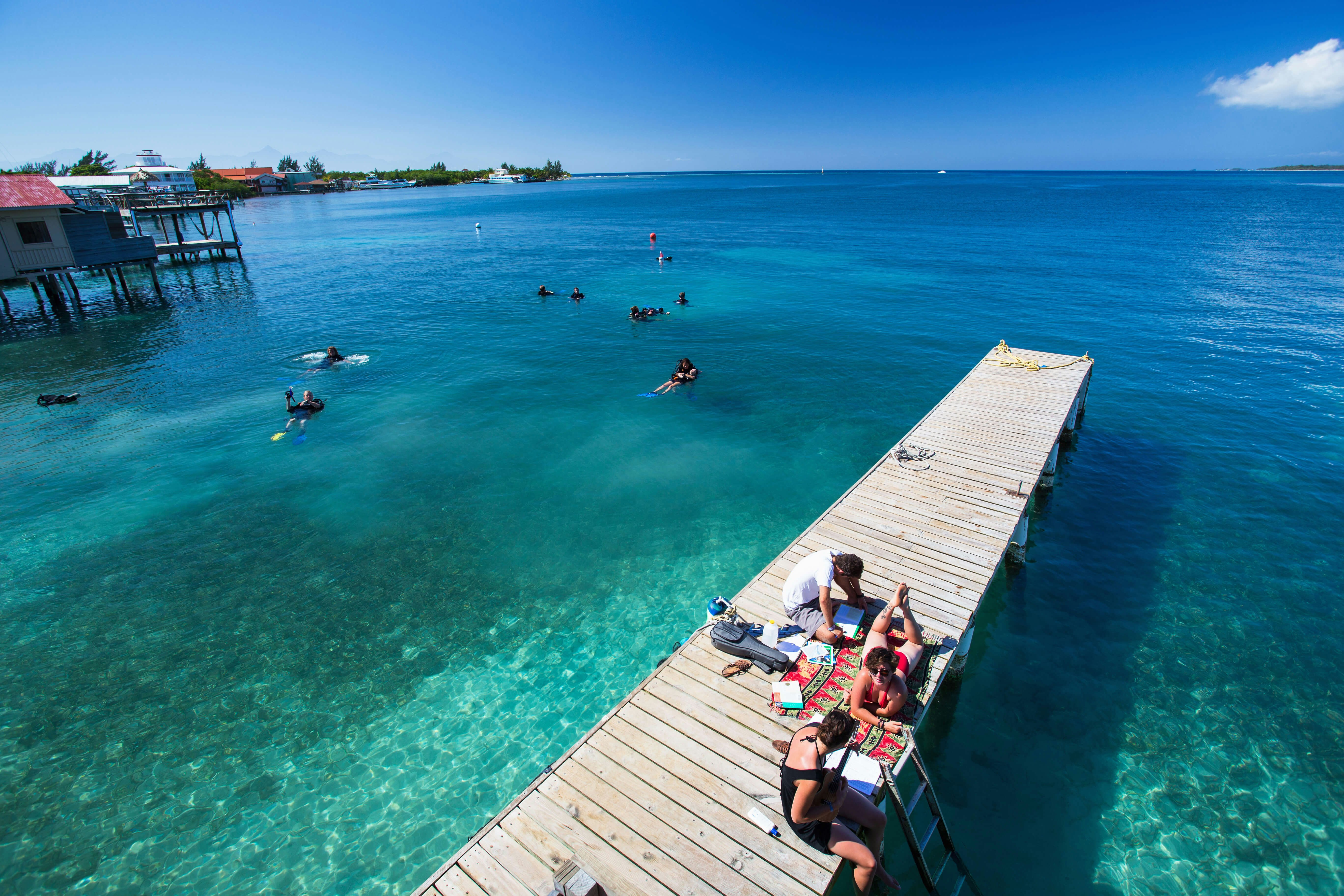 Girls play music in the sun on a Utila dock with snorkelers in the water behind them