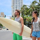Honolulu Hawaii lifestyle surfers people walking in city with surfboards going to the beach surfing. Outside Hawaiian surf living. Surfer couple crossing street. Waikiki, Honolulu, Oahu, Hawaii, USA.