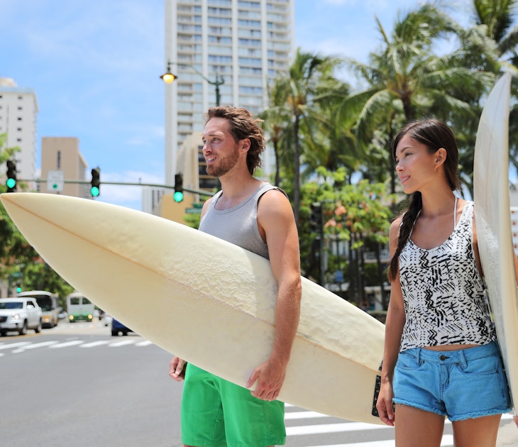 Honolulu Hawaii lifestyle surfers people walking in city with surfboards going to the beach surfing. Outside Hawaiian surf living. Surfer couple crossing street. Waikiki, Honolulu, Oahu, Hawaii, USA.