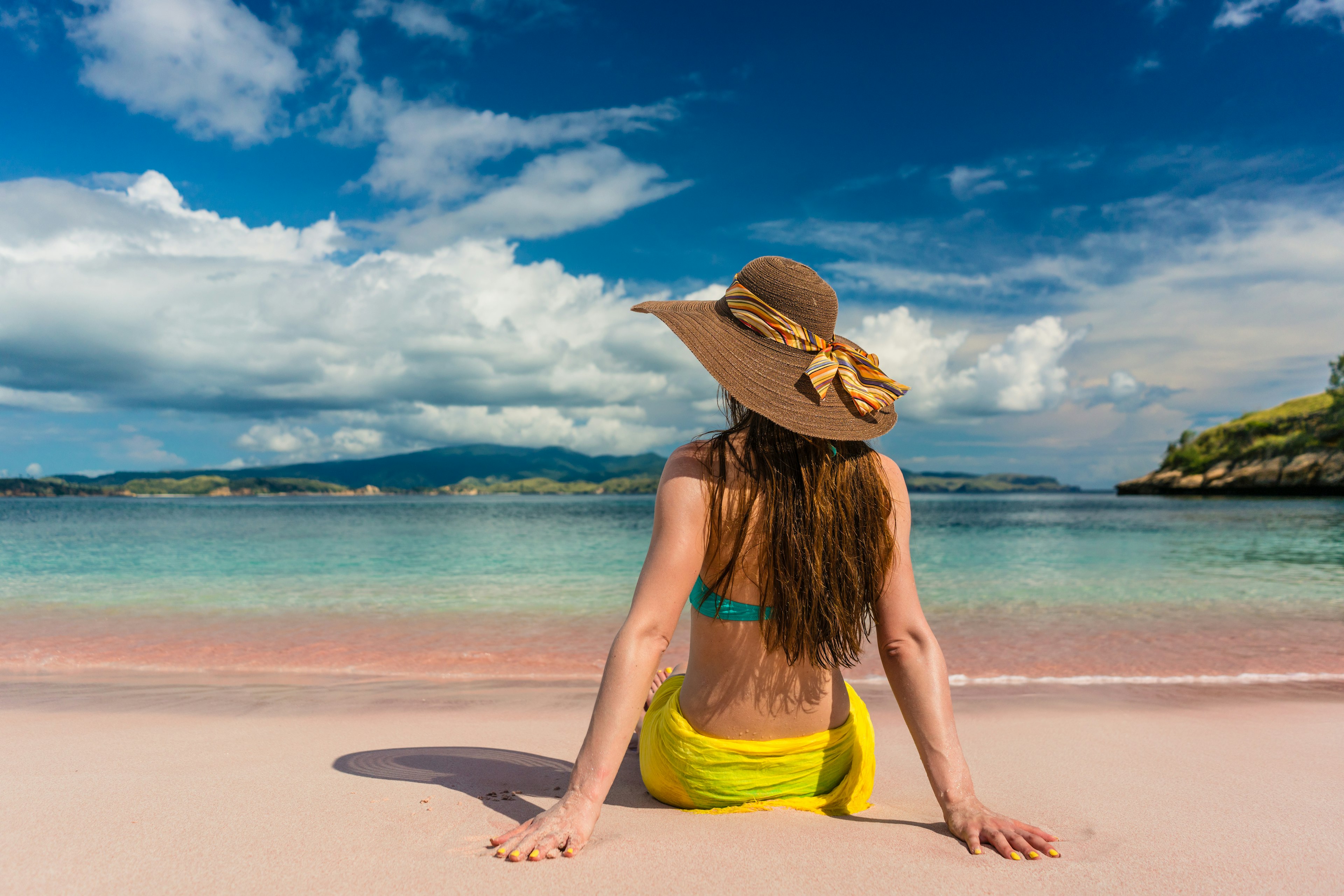 A young woman with straw hat sitting on the sand at Pink Beach, Komodo Island, Indonesia