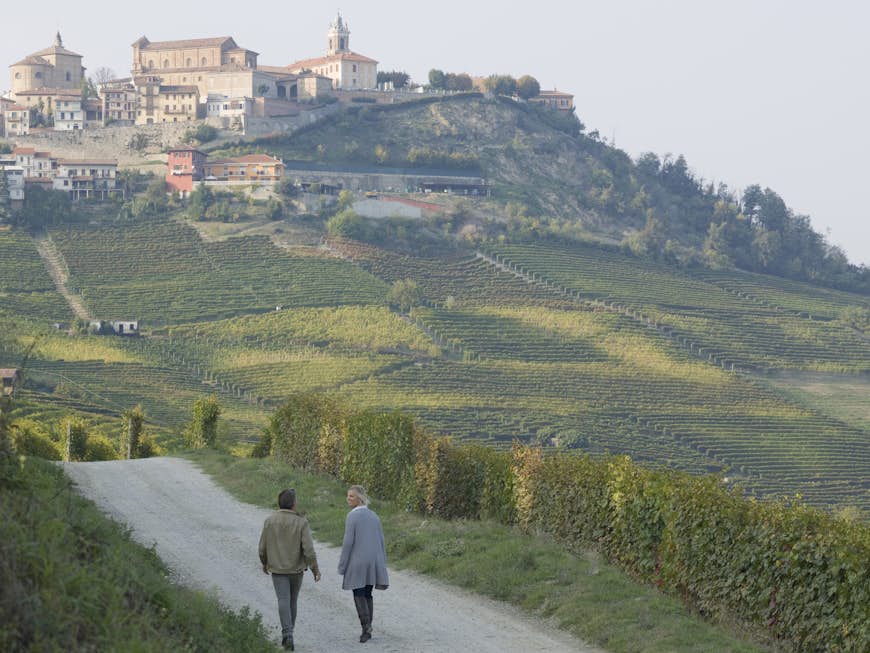 Par går förbi vingårdar längs en landsväg med den historiska byn La Morra i fjärran, Piemonte, Italien