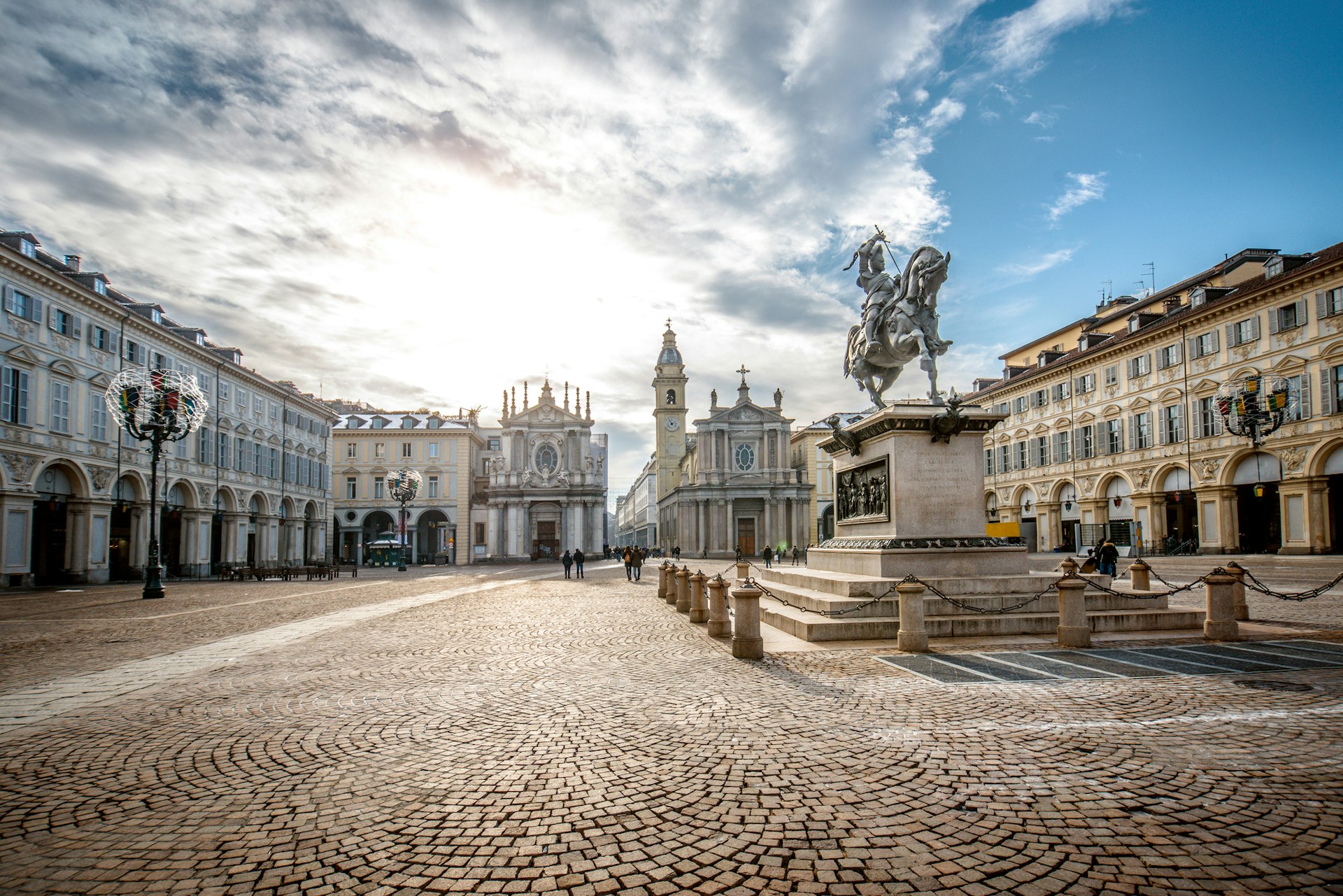 People walk through a large elegant cobbled square with two churches at one end