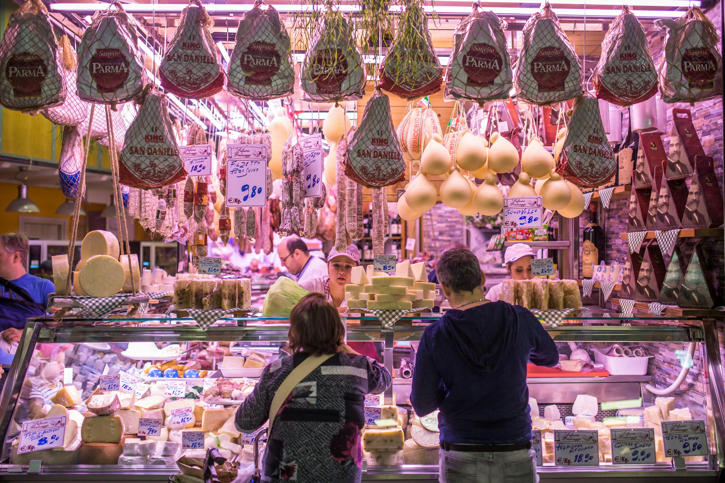 Two people stand at a cheese and meat counter in a market ordering food
