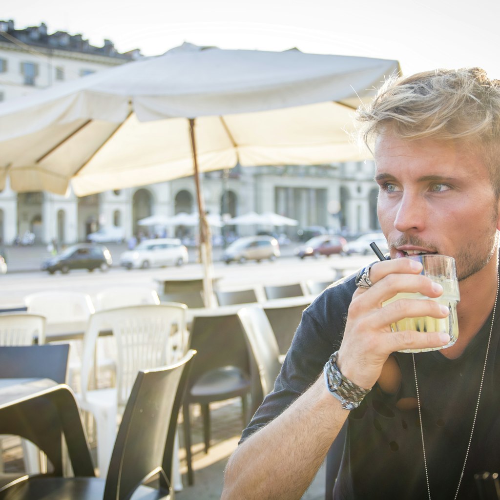 Handsome blonde young man sitting in Turin cafe holding glass wearing black t-shirt looking away.