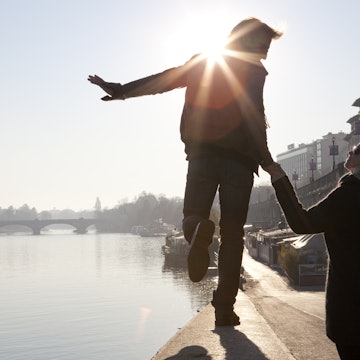 A man walks along a riverside stone wall in Turin during the late afternoon with his partner, a woman, holding his hand as he looks towards her.