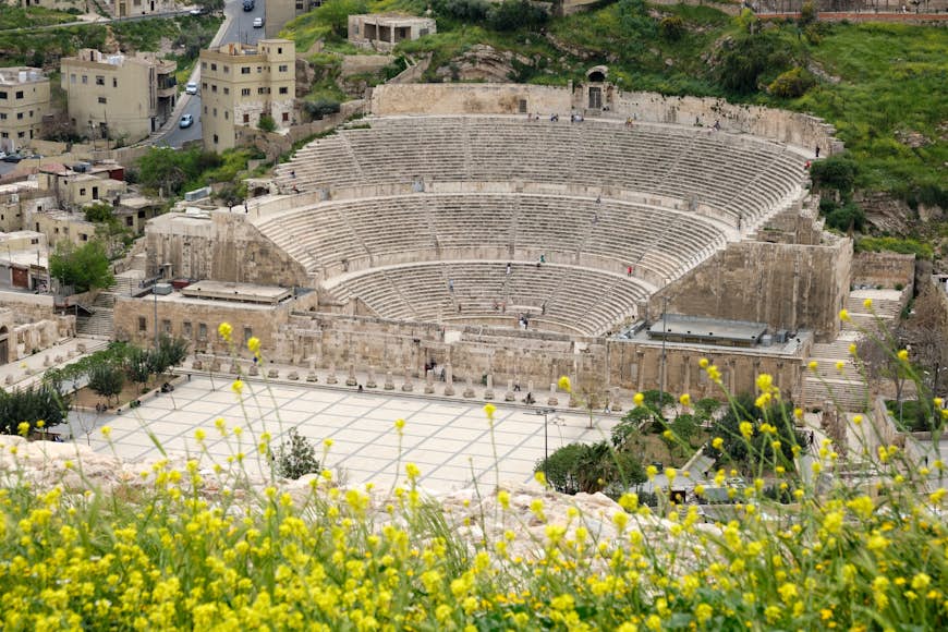 A vast Roman amphitheater is toured by visitors while yellow spring flowers are in bloom on the hillside above