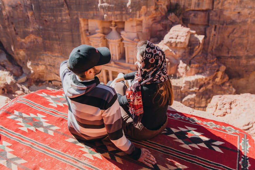 A couple of travelers sit on a red blanket on a cliff above the ancient city of Petra, carved into orange rock