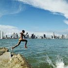 Boy jumping of rock into water in Casco Viejo bay with Panama City in background.