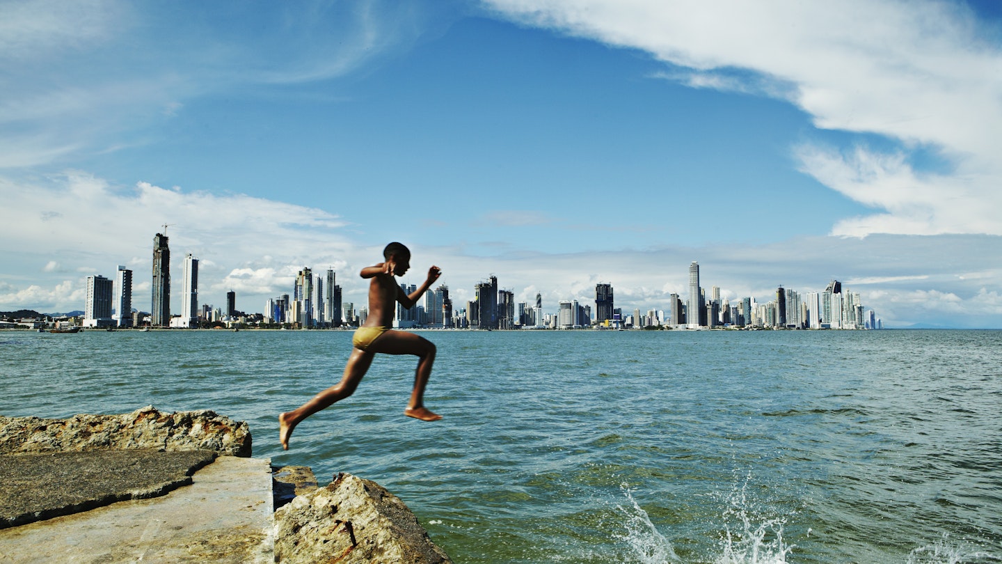 Boy jumping of rock into water in Casco Viejo bay with Panama City in background.