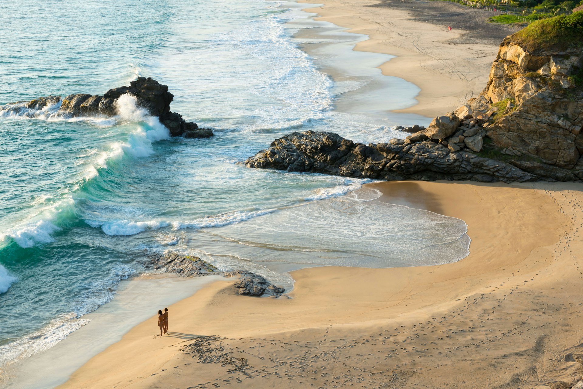 A man and woman face the Pacific Ocean while standing nude together on a wide sand beach in front of crashing waves 