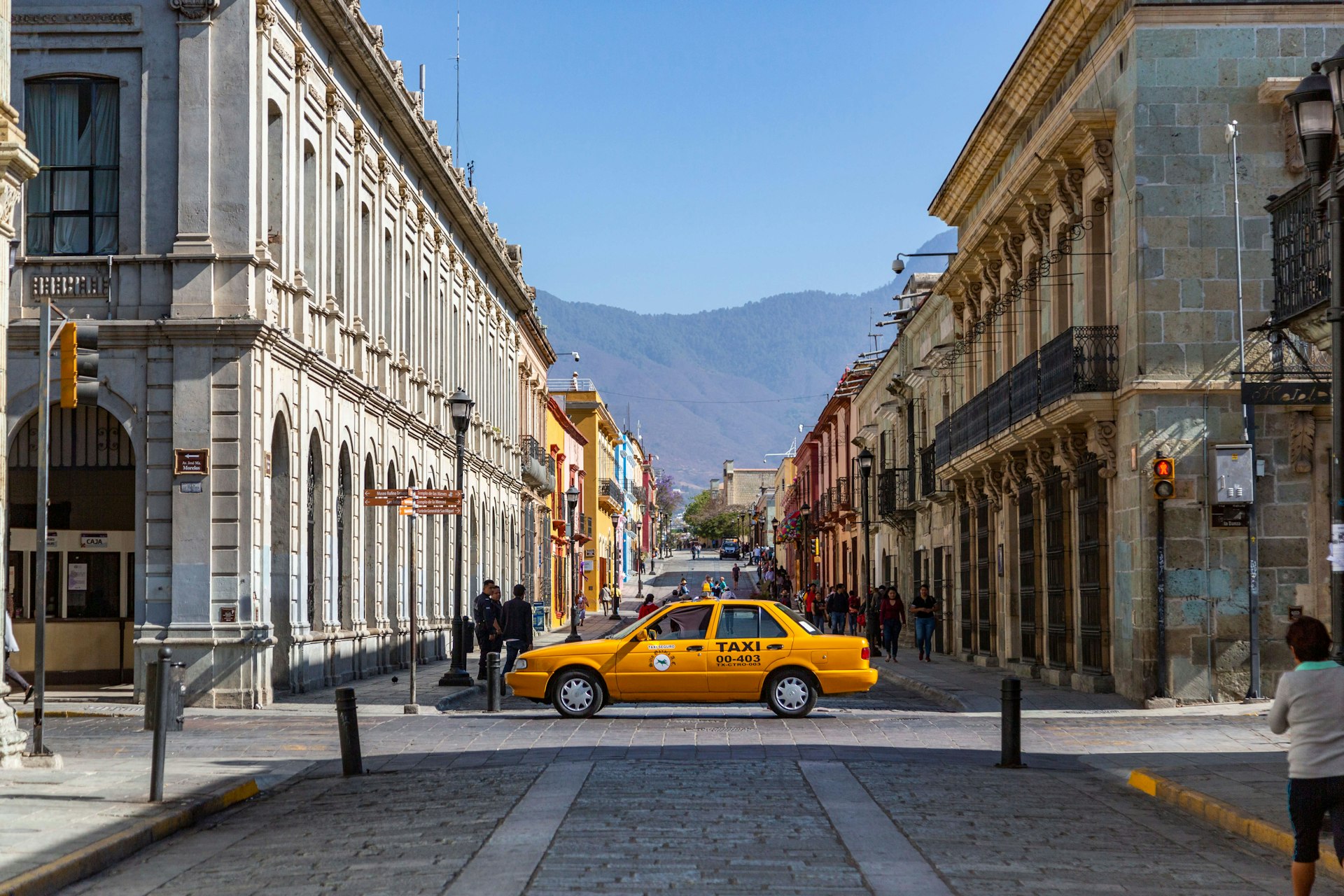 A sunny morning in Oaxaca - a yellow taxi in the center of a big avenue with some people walking by 