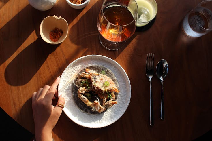 An overhead table shot with a drinking glass and a hand next to a plate holding a deep-fried soft-shell crab on a blue-corn tortilla 