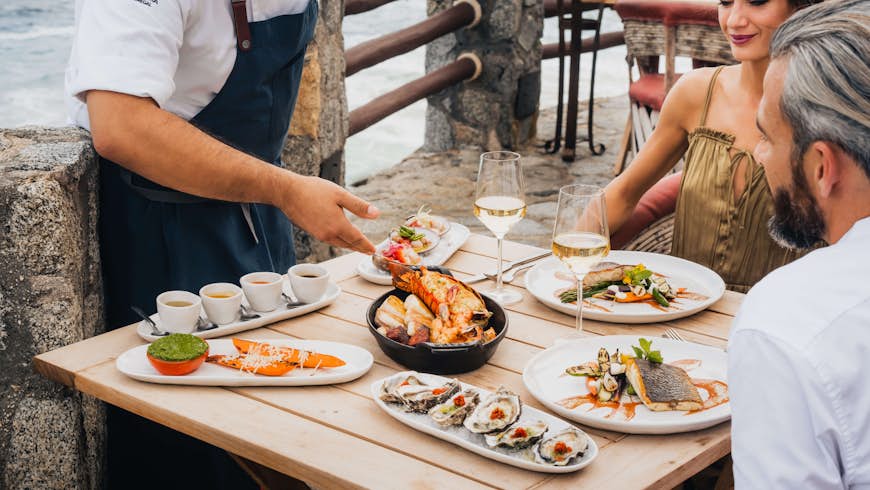 Two diners sitting at a table full of food on Pedregal's Champagne Terrace as a waiter presents the dishes