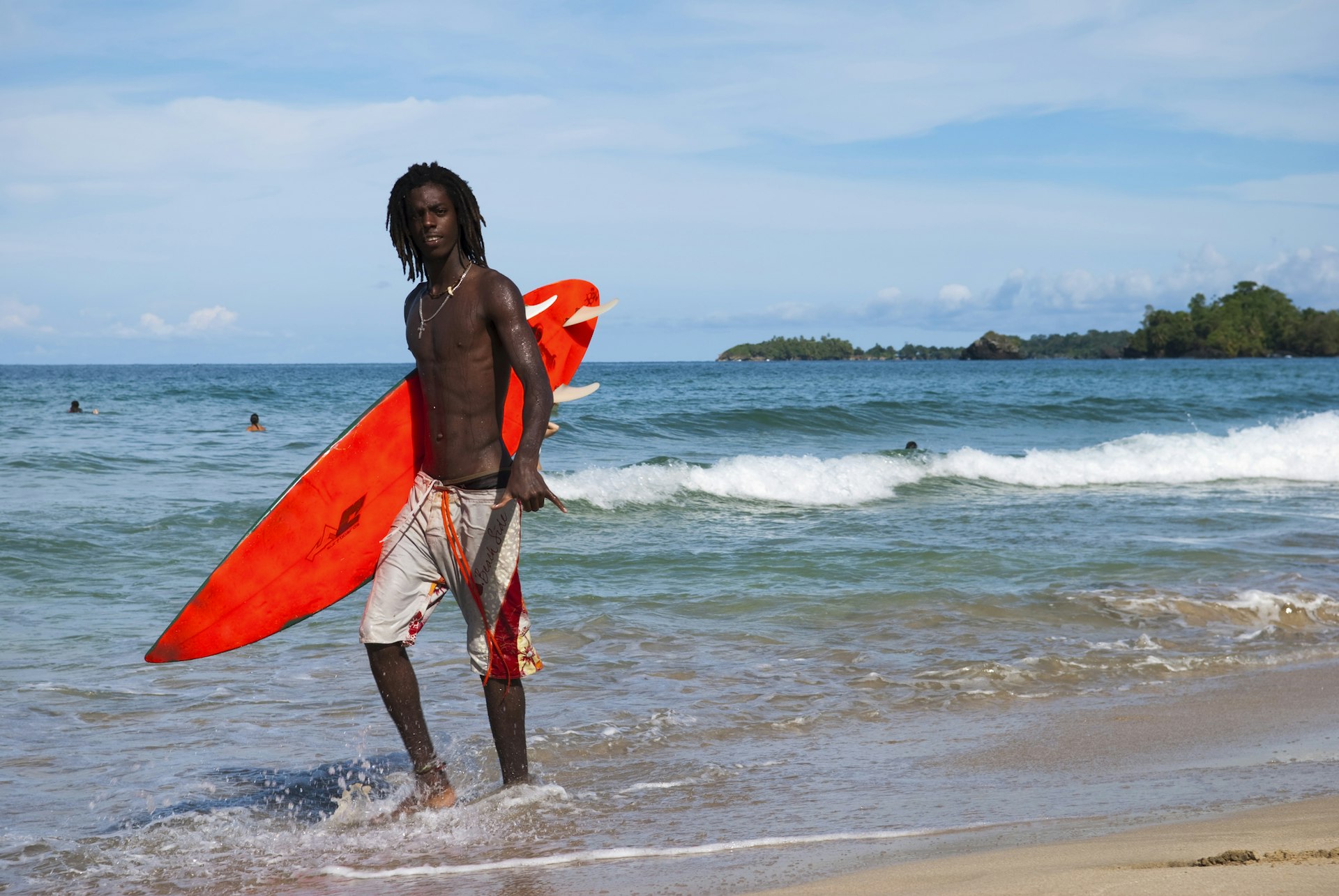 A Panamanian surfer with dread locks walks with his surfboard at Wizard Beach, located on Isla Bastimentos in the Bocas del Toro Archipelago. He is giving the hang loose sign with his left hand.