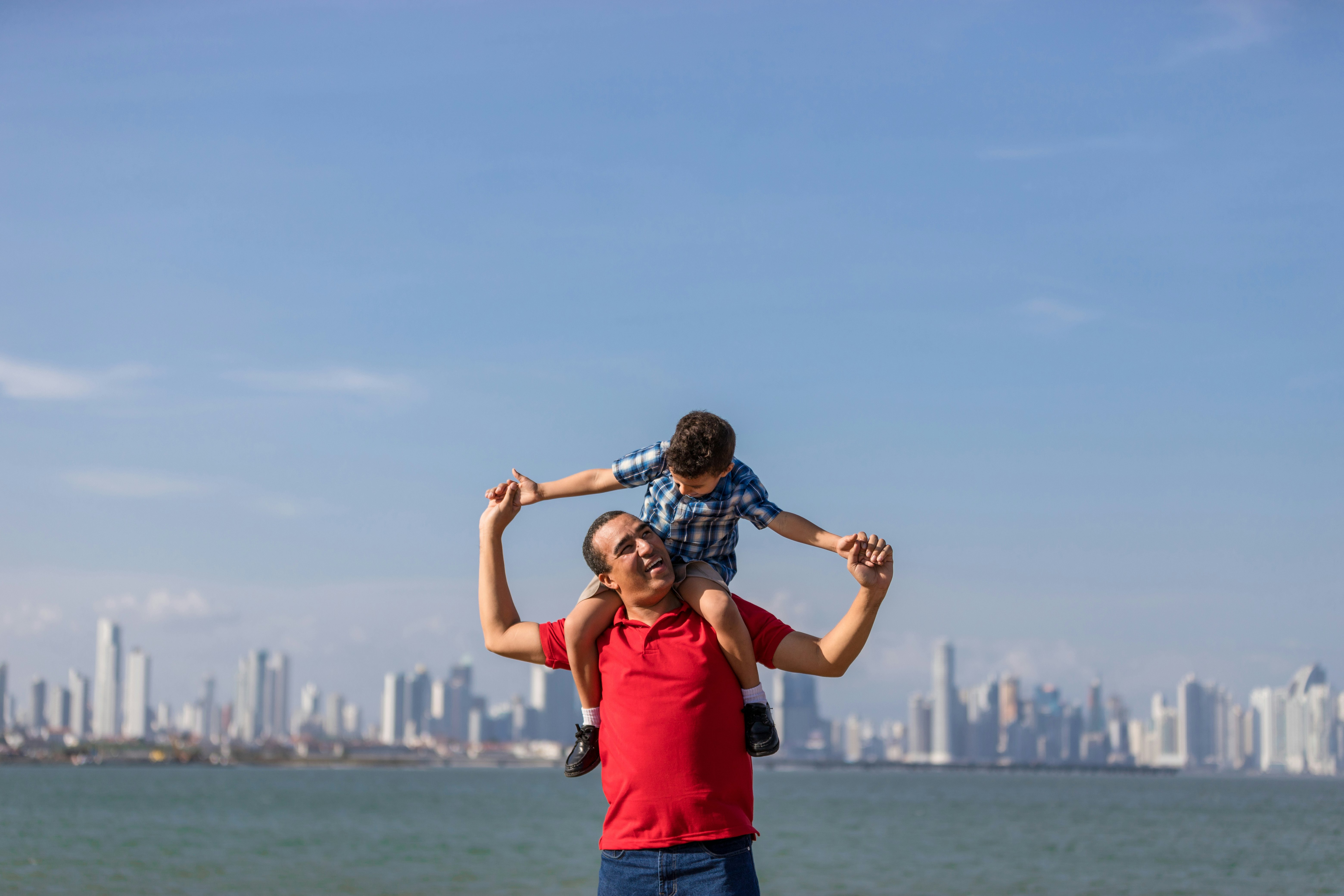 A young boy sits on a man's shoulders at the edge of a bay lined with the high-rise buildings of Panama City