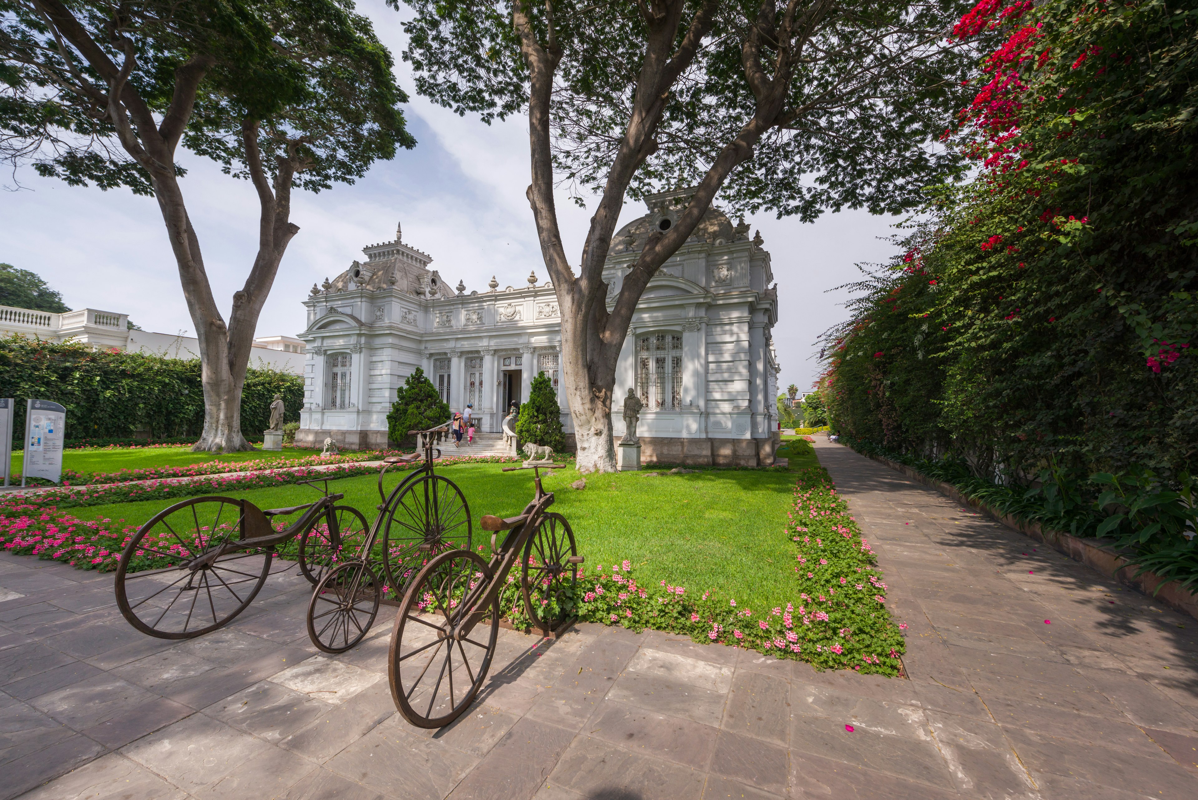 A grand white building with lush gardens and a sculpture of two bicycles as if they are parked up outside