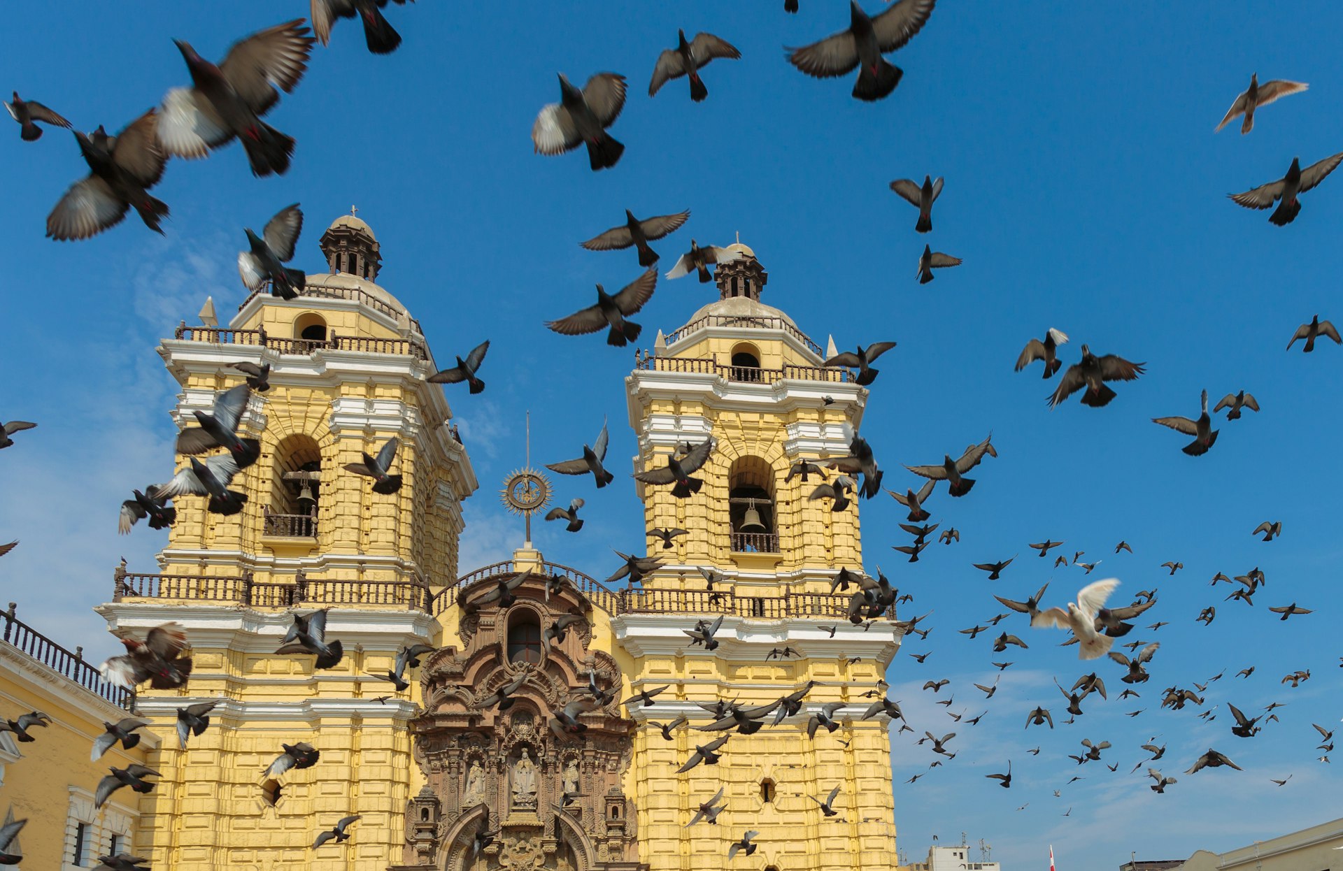 A flock of pigeons fly upwards into a blue sky above a yellow church building with two prominent bell towers