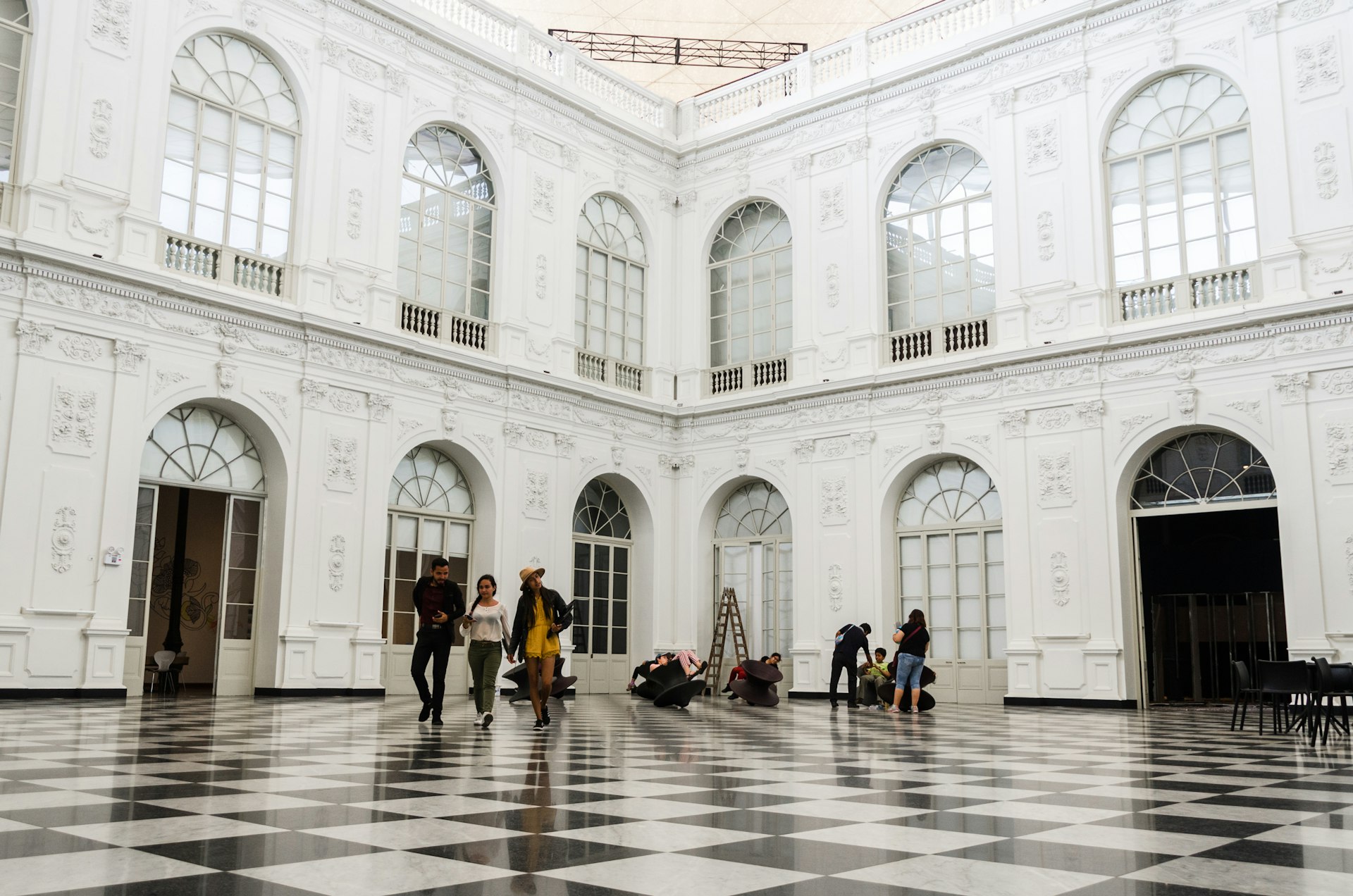 People walk across a vast black-and-white checked floor surrounded by white walls with arched windows