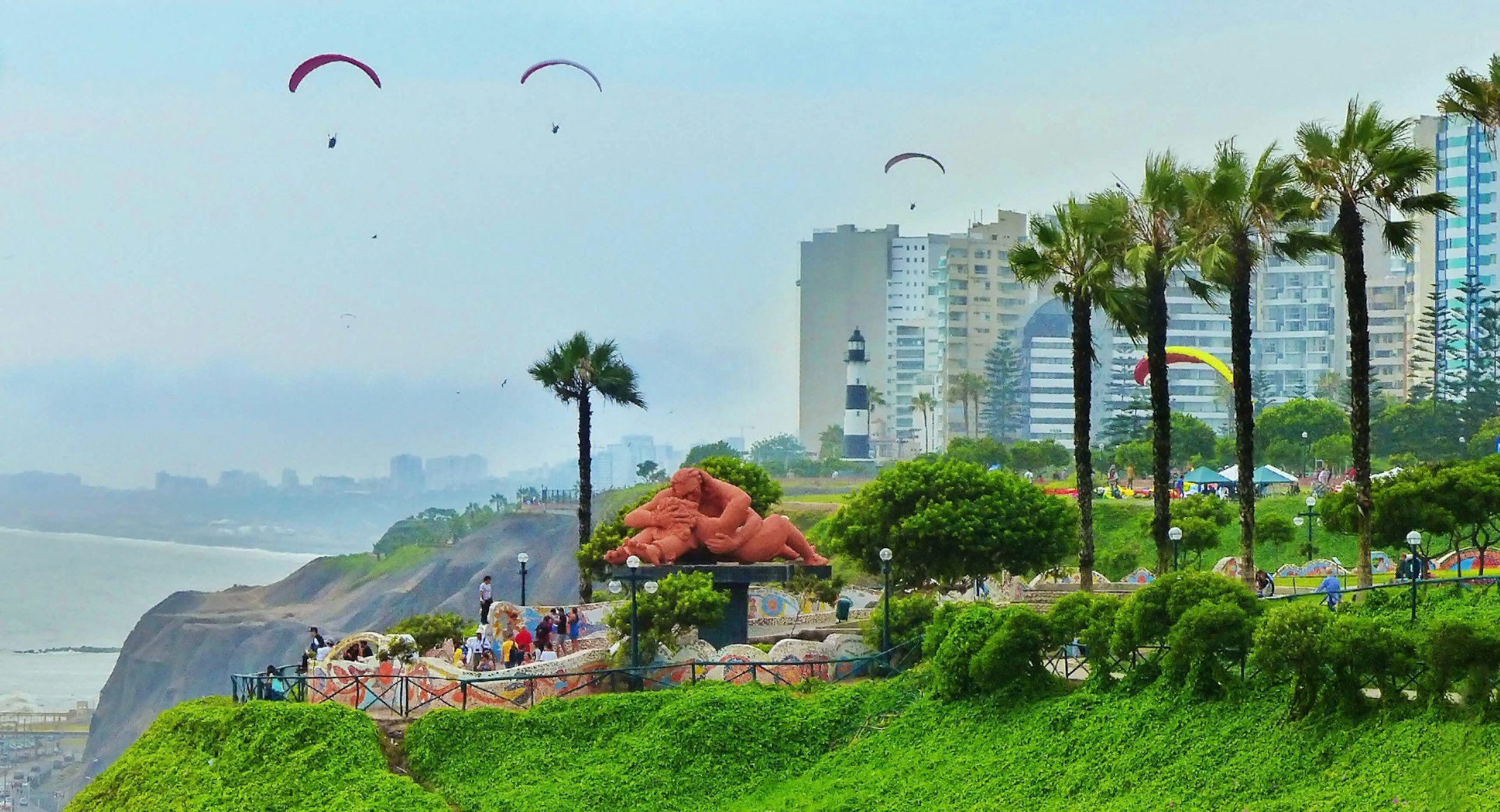 People in a park on a coastal cliff overlooking the Pacific Ocean, with a red statue of a couple embracing, paragliders overhead and a lighthouse in the background