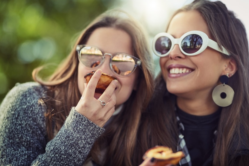 Two young women, each wearing sunglasses, eat small peach pies while sitting in the park. 