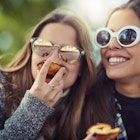 Two young women, each wearing sunglasses, eat small peach pies while sitting in the park. 