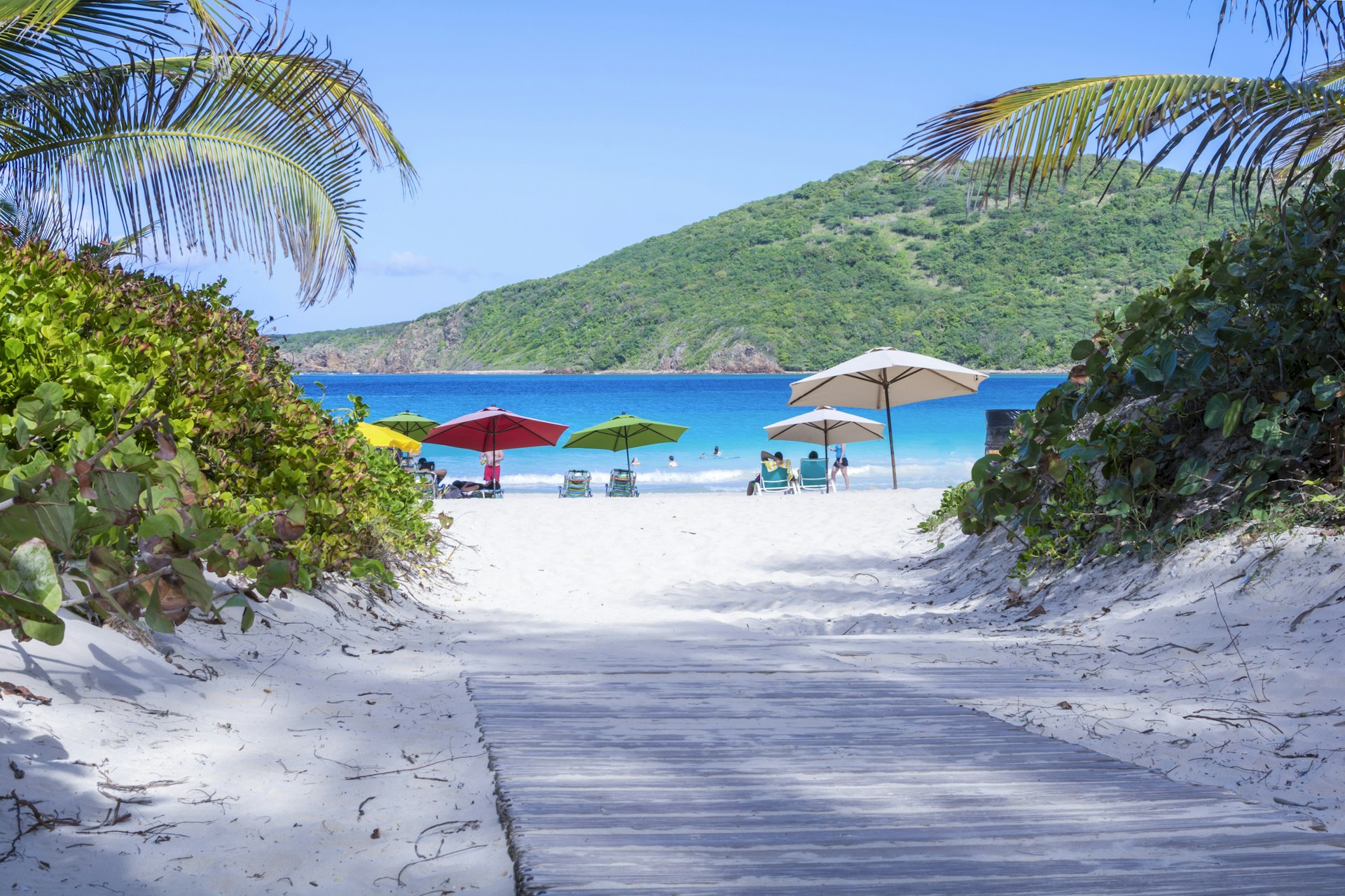 A wooden boardwalk dusted with white sand leads down to a beach where people relax under colorful umbrellas on the island of Culebra, Puerto Rico