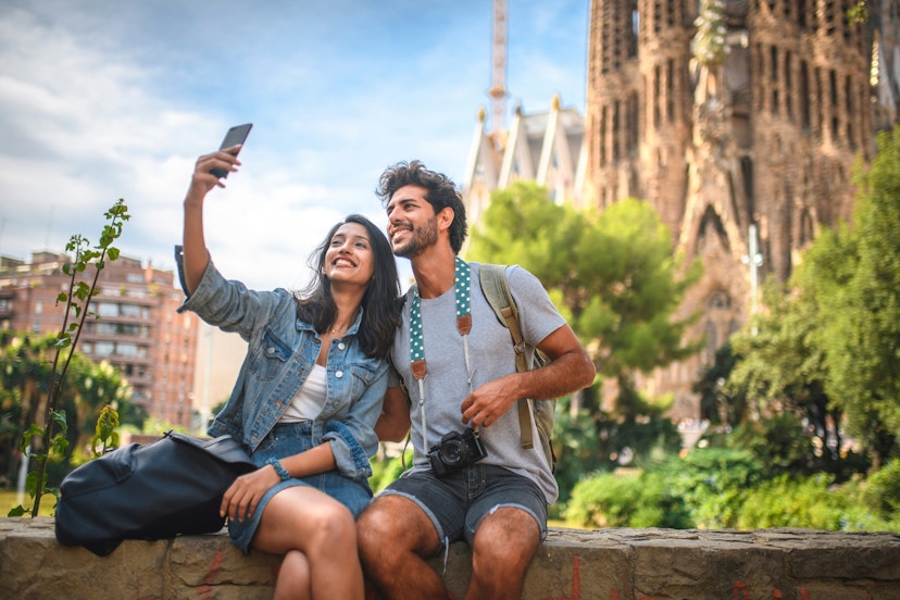 Male and female travelers sitting on wall in public park near Sagrada Familia in Barcelona and taking selfie on sunny summer day.