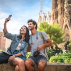 Male and female travelers sitting on wall in public park near Sagrada Familia in Barcelona and taking selfie on sunny summer day.