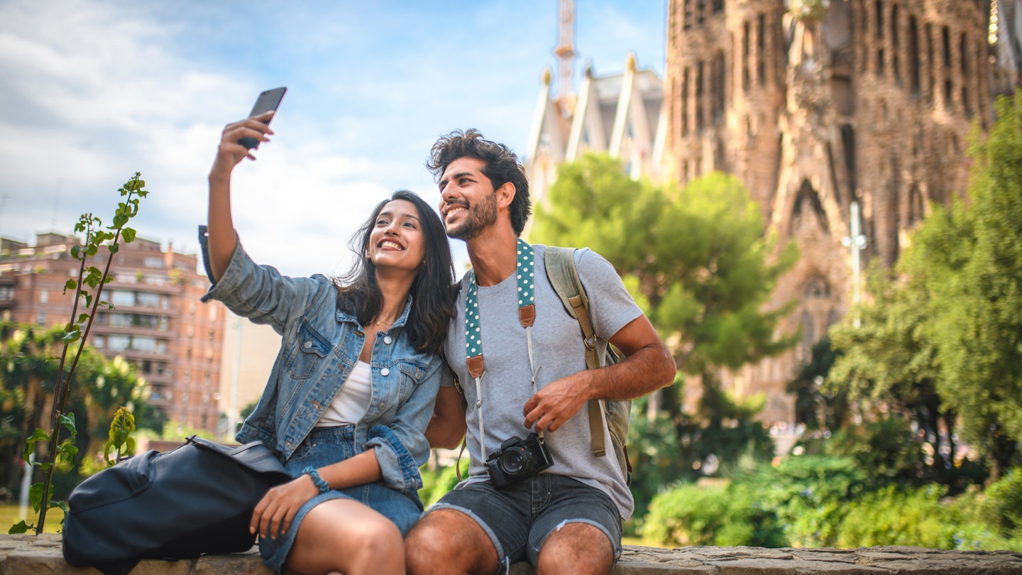 Male and female travelers sitting on wall in public park near Sagrada Familia in Barcelona and taking selfie on sunny summer day.