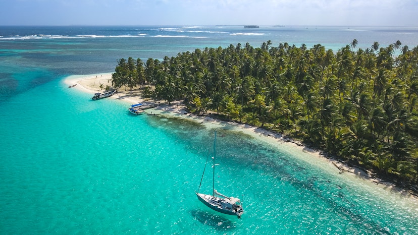 Aerial drone shot of a sailing yacht anchored in turquoise water right next to a white sand beach of remote tropical island full of green palm trees in Caribbean Sea.