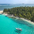 Aerial drone shot of a sailing yacht anchored in turquoise water right next to a white sand beach of remote tropical island full of green palm trees in Caribbean Sea.