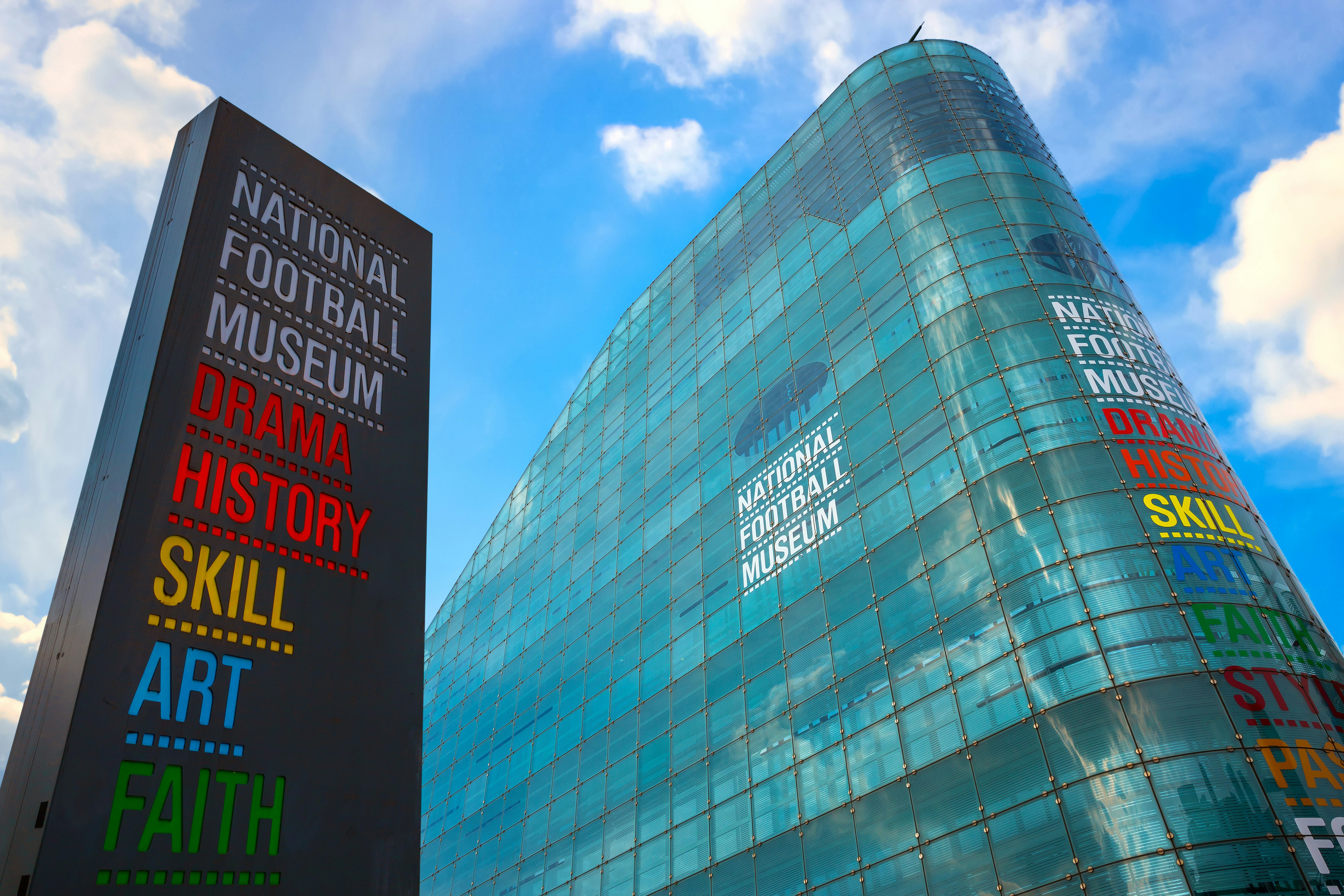 A tall curved glass building with a large sign outside it. The colorful writing says "National Football Museum: Drama, History, Art, Skill, Faith
