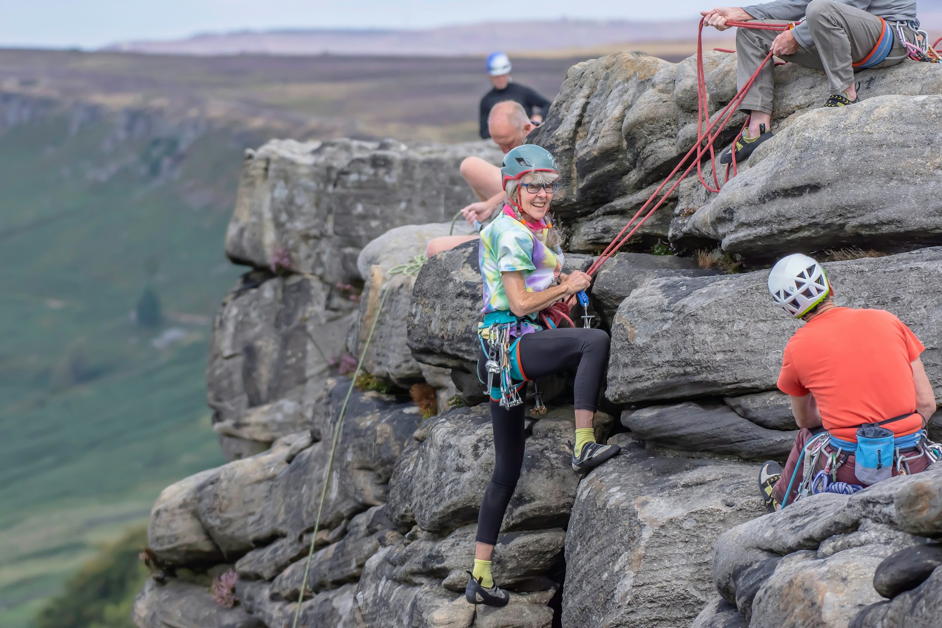 A lady in her 60s wearing climbing gear and ropes smiles as she attempts to climb down a cliff face
