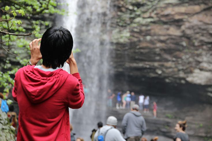 A person wearing a red jacket seen from behind, with a bunch of people in front of a waterfall blurred in the background