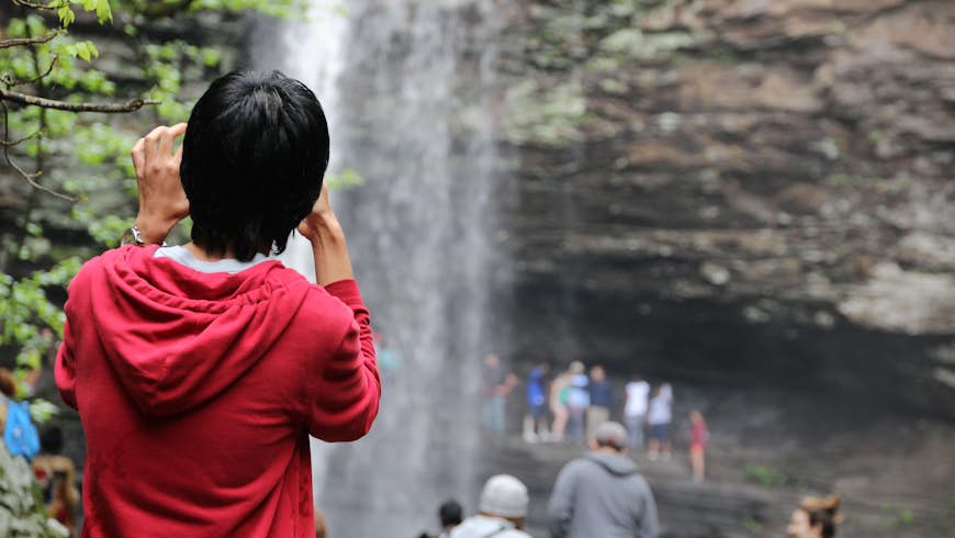 A person wearing a red jacket seen from behind, with a bunch of people in front of a waterfall blurred in the background