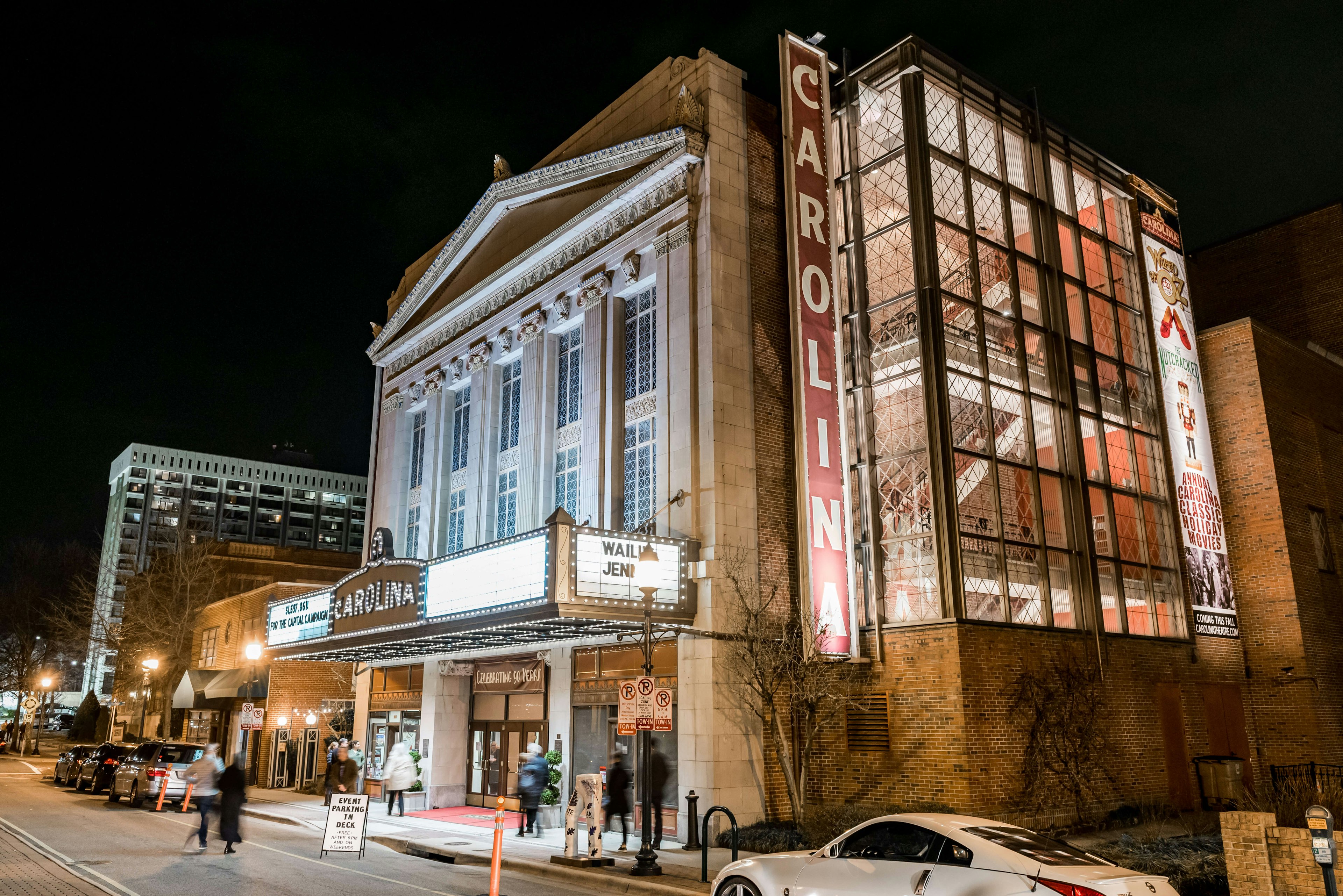 People walking and cars parked in front of The Carolina Theatre in downtown Greensboro with marquee lights lit up at night.