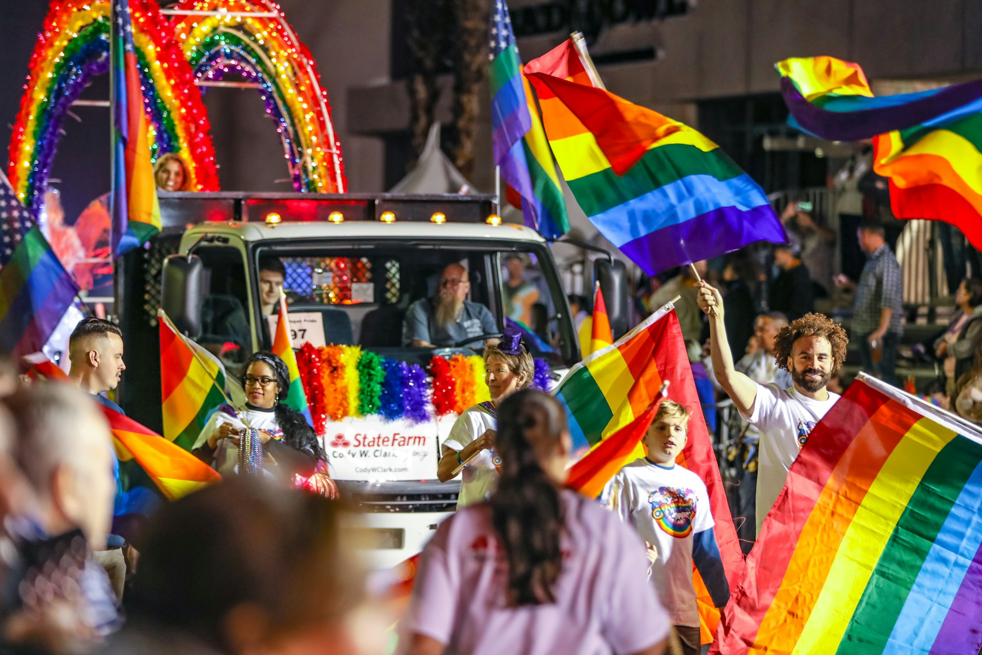 People take part in a parade walking in front of a low-bed truck. There are rainbow flags and motifs flying everywhere