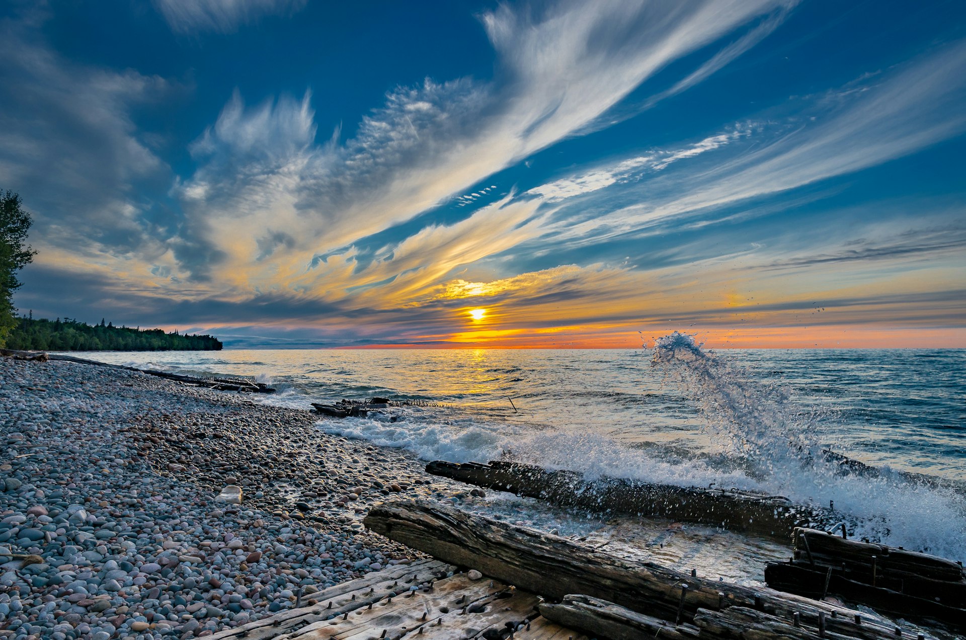 The sun sets over the lake; the shore is littered with driftwood