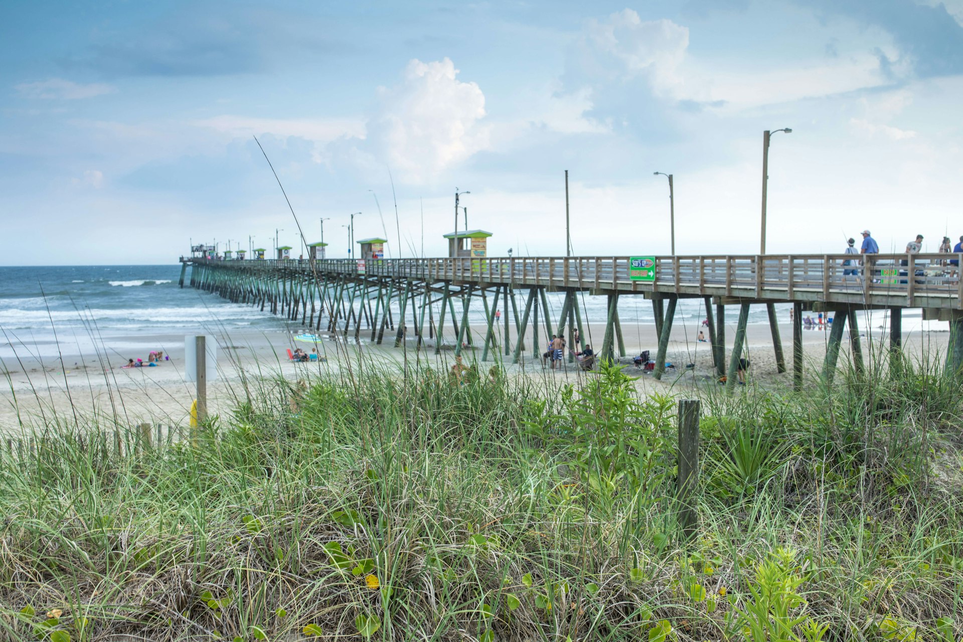 A long wooden fishing pier, seen from behind the dunes with grass in the foreground