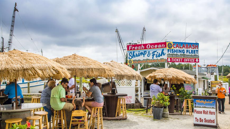 Patrons eating dockside at high tables with thatched umbrellas at Waterfront Seafood Shack