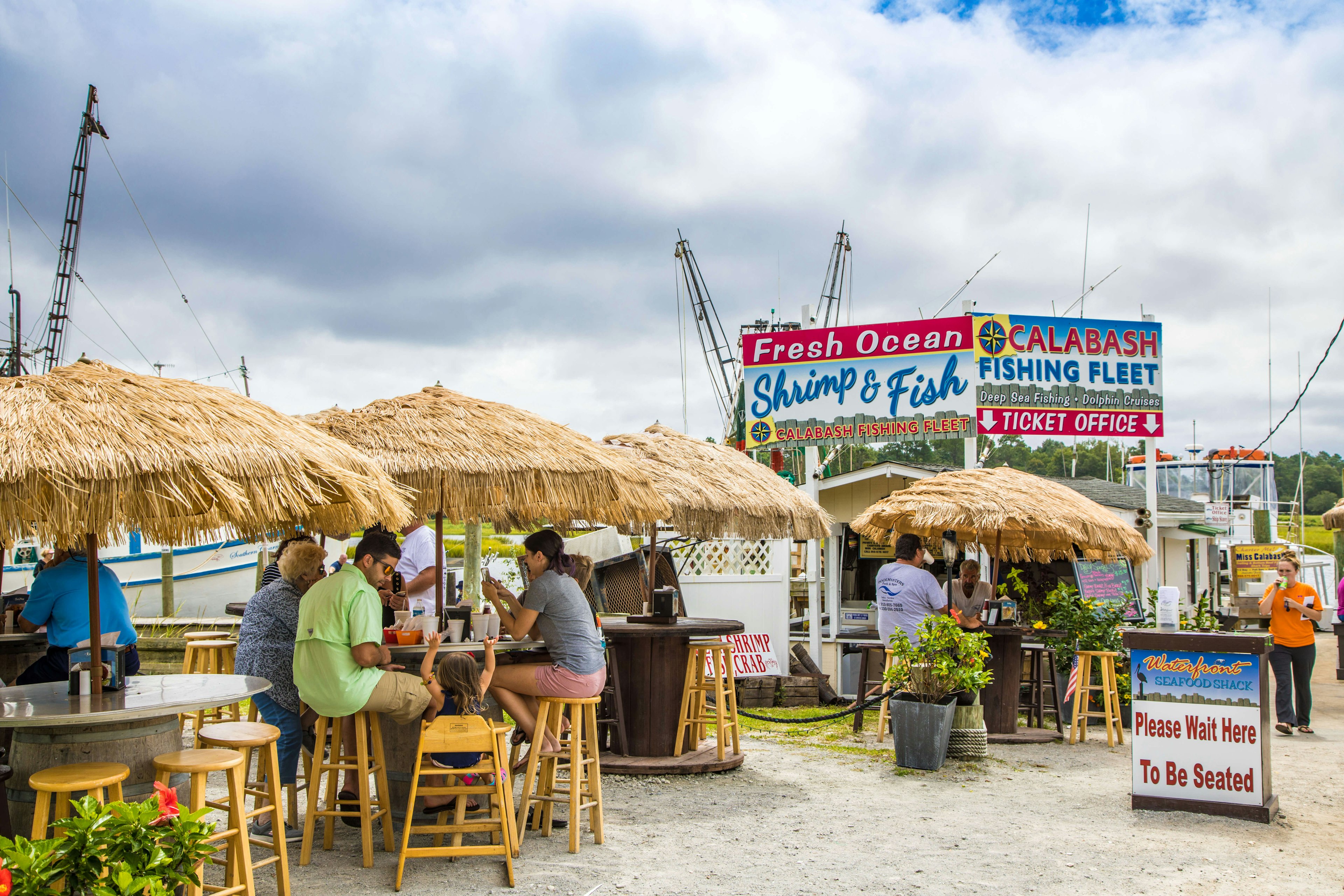 Patrons eating dockside at high tables with thatched umbrellas at Waterfront Seafood Shack